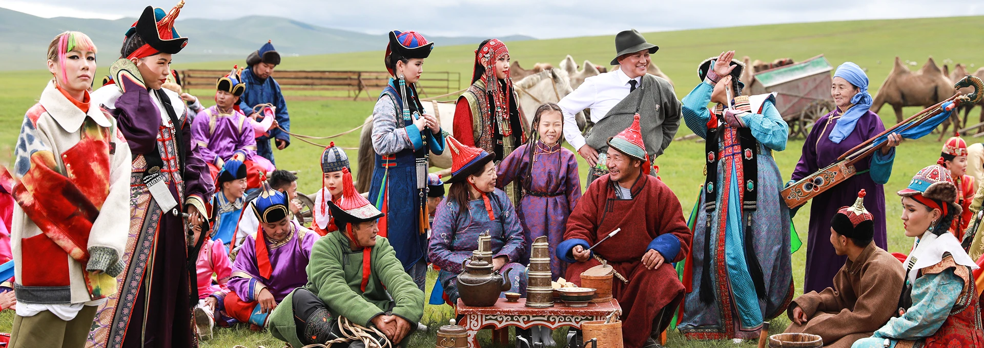 Mongolian people in traditional dress around a snack table on grassland, with camels and horses behind