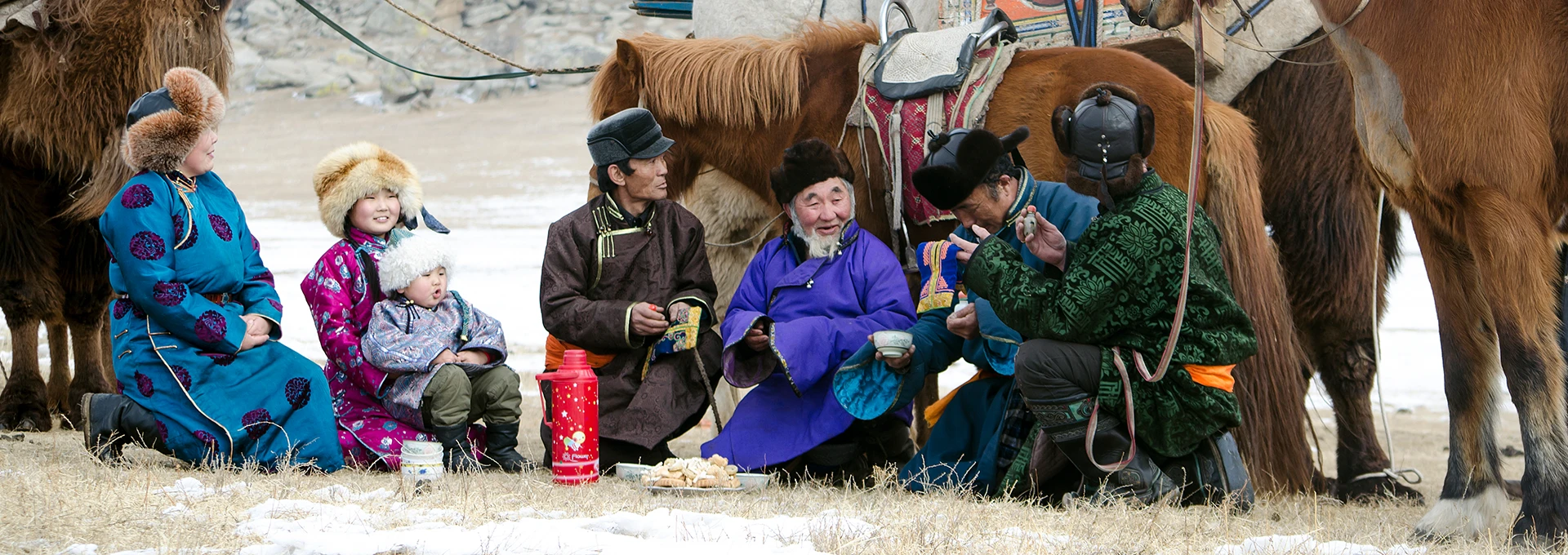 Mongolian family with horses in the back eating food on a grass plane covered with some snow